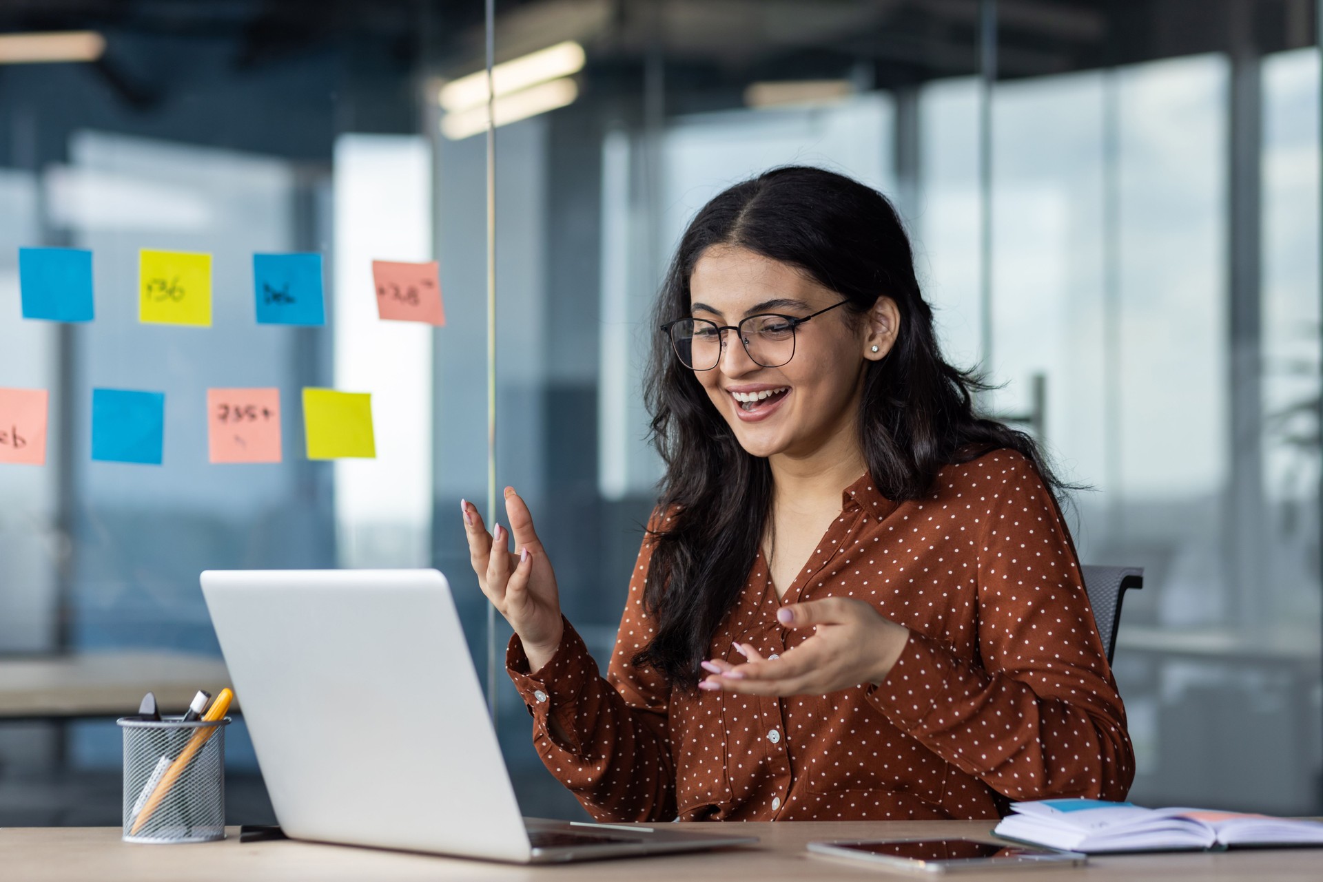 Young business woman inside office using laptop for video call, female worker smiling and talking with colleagues remotely in online meeting, joyful female worker with computer